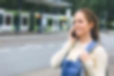 Junge Frau mit Blaumannhose wartet telefonierend an einer Haltestelle.